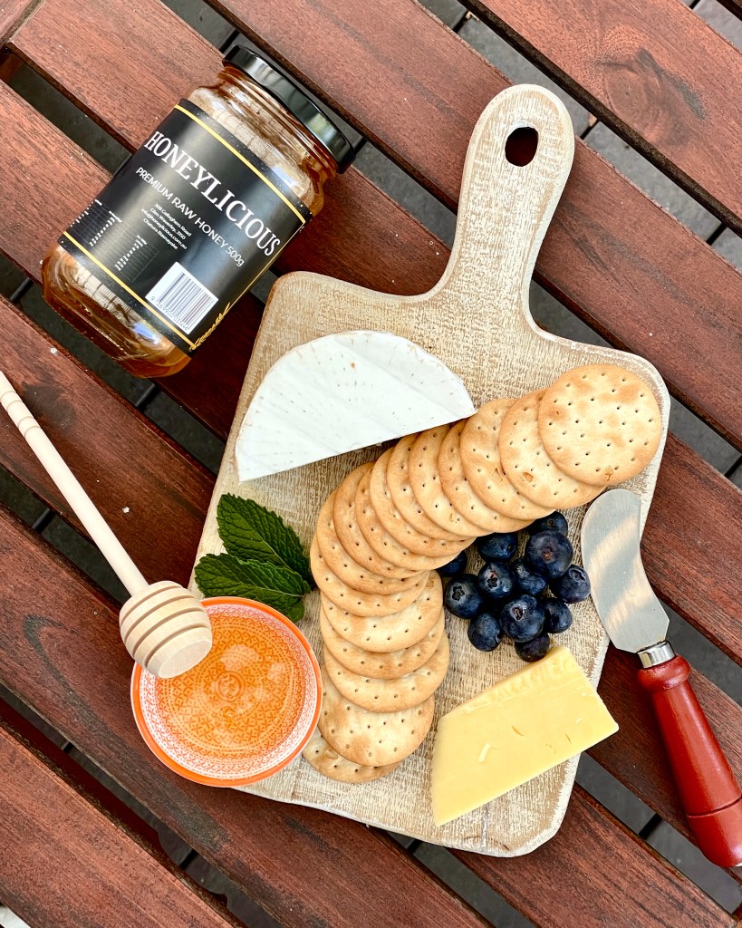 Flatlay of a cheese board with brie, cheddar, crackers, bluberries and Honeylicious honey
