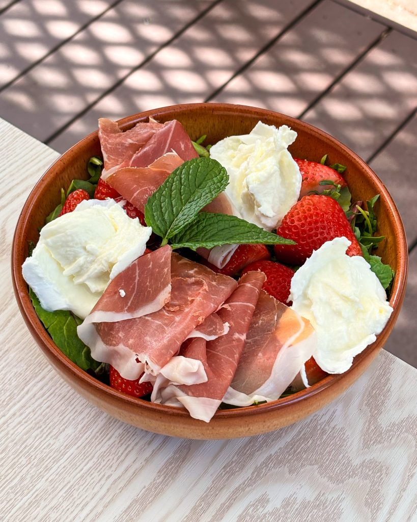 Brown bowl on off white and brown background. Bowl contains mozzarella cheese, bitter leaves, prosciutto, strawberries and mint