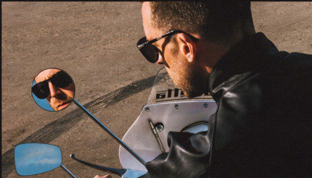 Miles Kane with his back to camera sat on a motorcycle. His face can be seen reflected in the wing mirror.