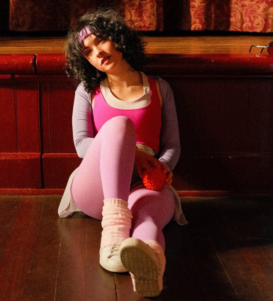 Singer Aleksiah in an 80s inspired pink acrobatic outfit sat on a wooden floor 