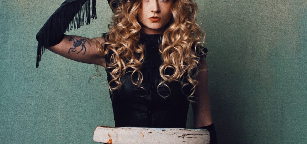 Country singer Janet Devlin in a black outfit against a grey background with her hand on her head