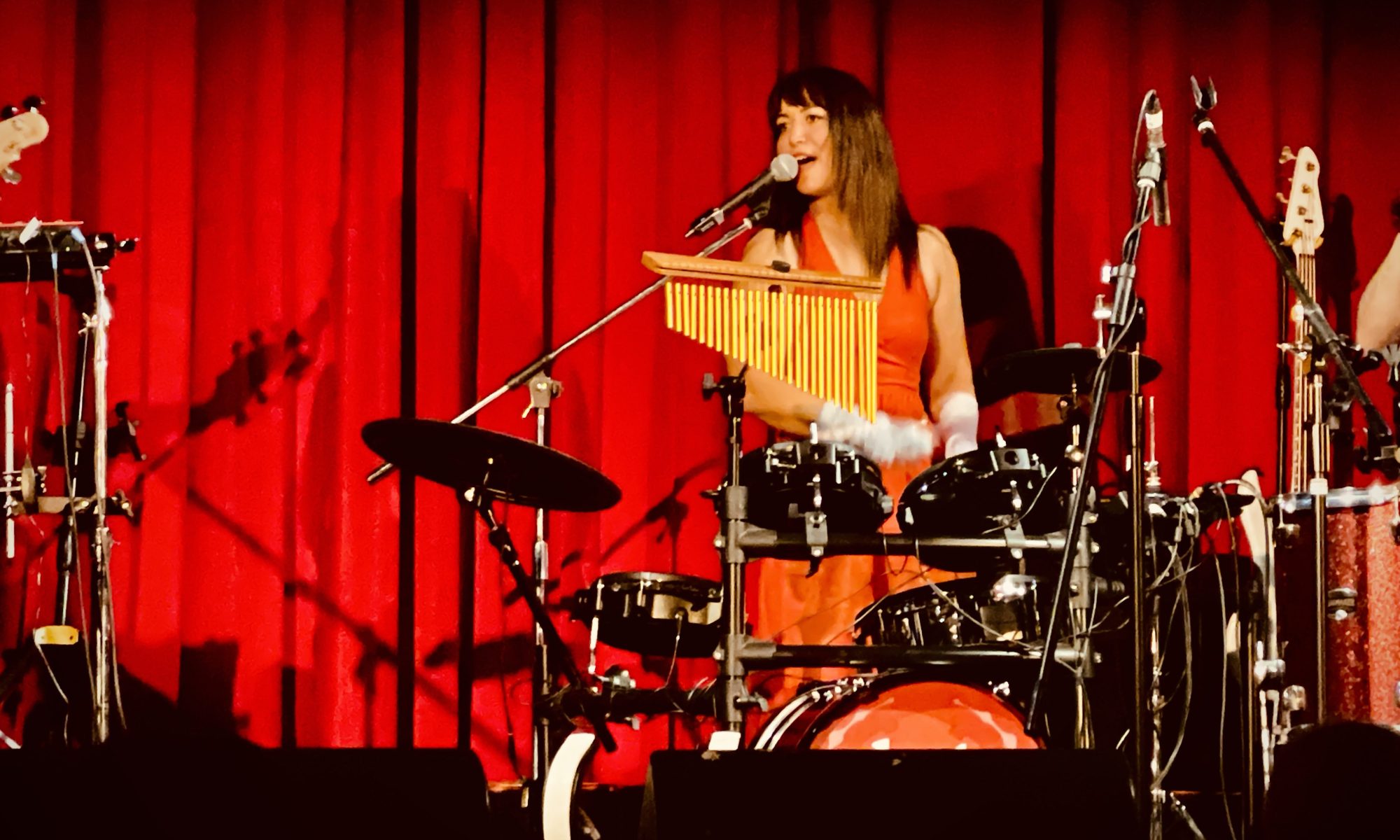 Musician Kat Greta playing the drums against a red background as a part of her Rhythm and Reverie tour