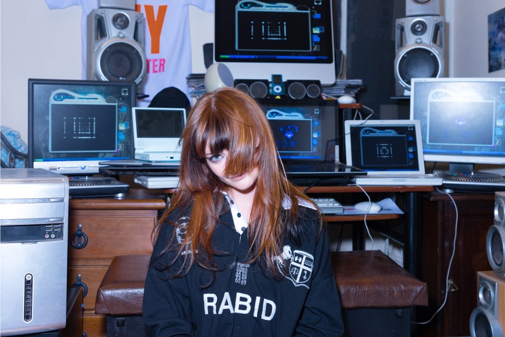 Ninajirachi with her hair convering her face in front of a desk full of computers.