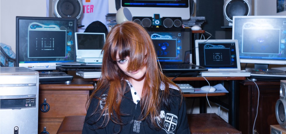 Ninajirachi with her hair convering her face in front of a desk full of computers.