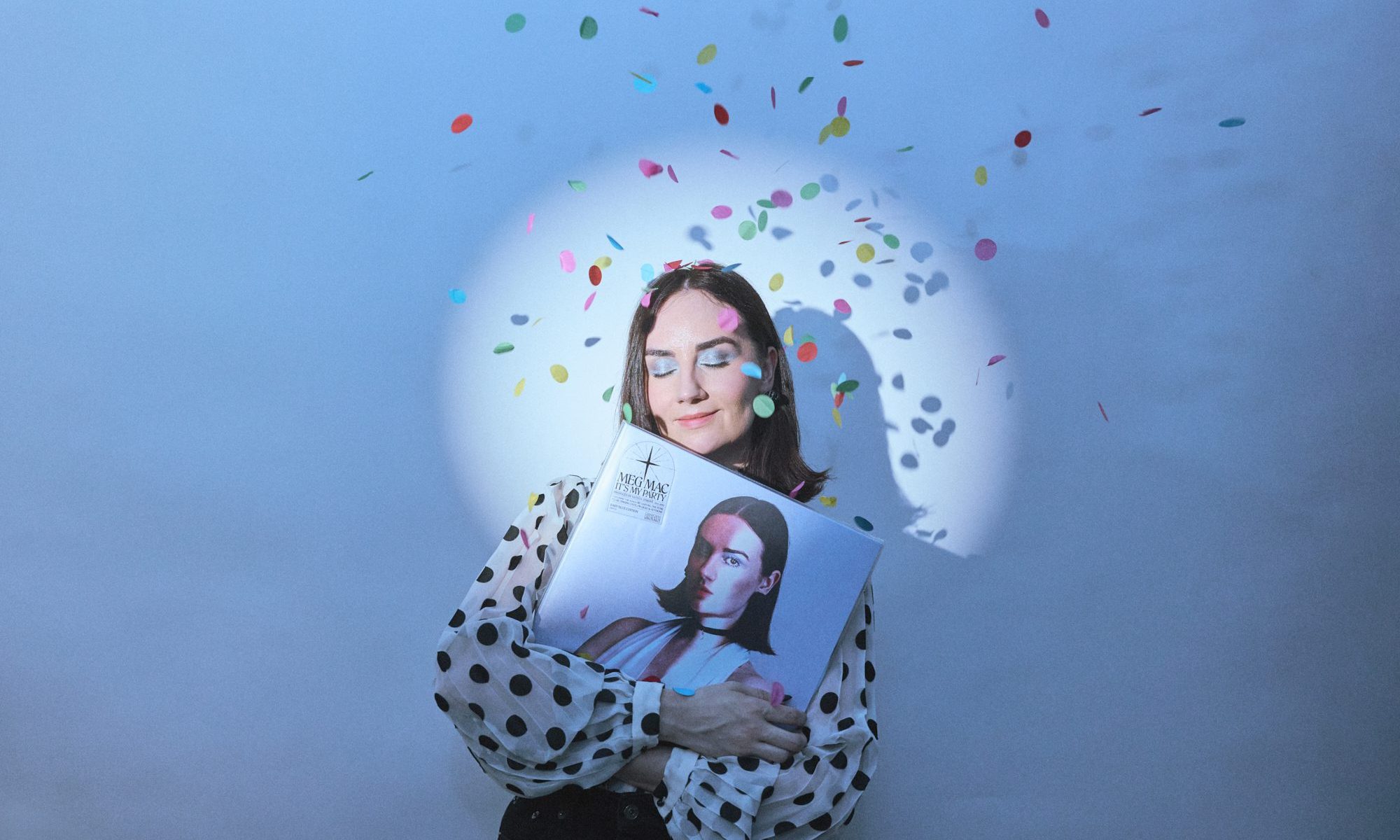 Musician Meg Mac holding her latest album on vinyl, with a spotlight on her face and confetti falling from the ceiling.