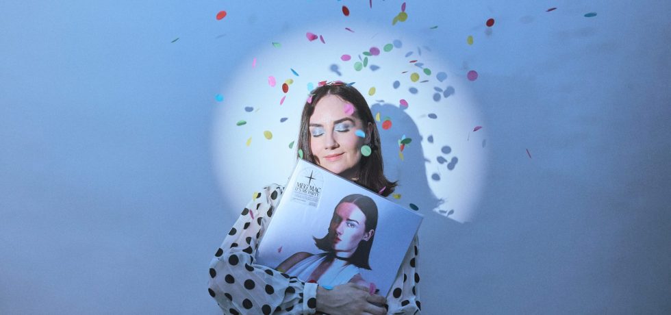 Musician Meg Mac holding her latest album on vinyl, with a spotlight on her face and confetti falling from the ceiling.