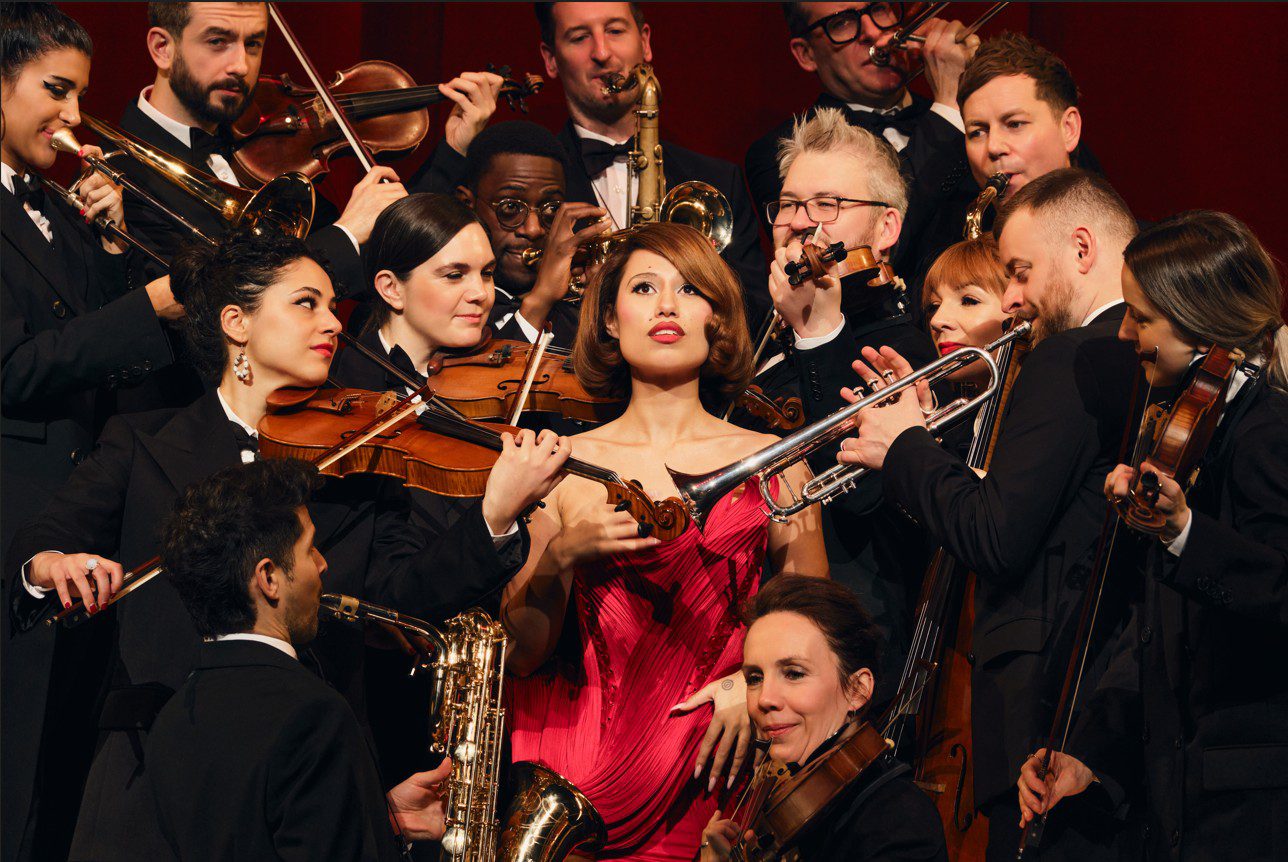 Image of RAYE in a red dress surrounded by an orchestra