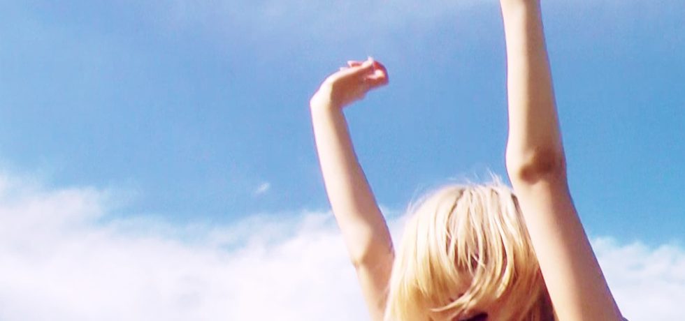 Image of Daine against a blue sky with clouds with their hands above their head