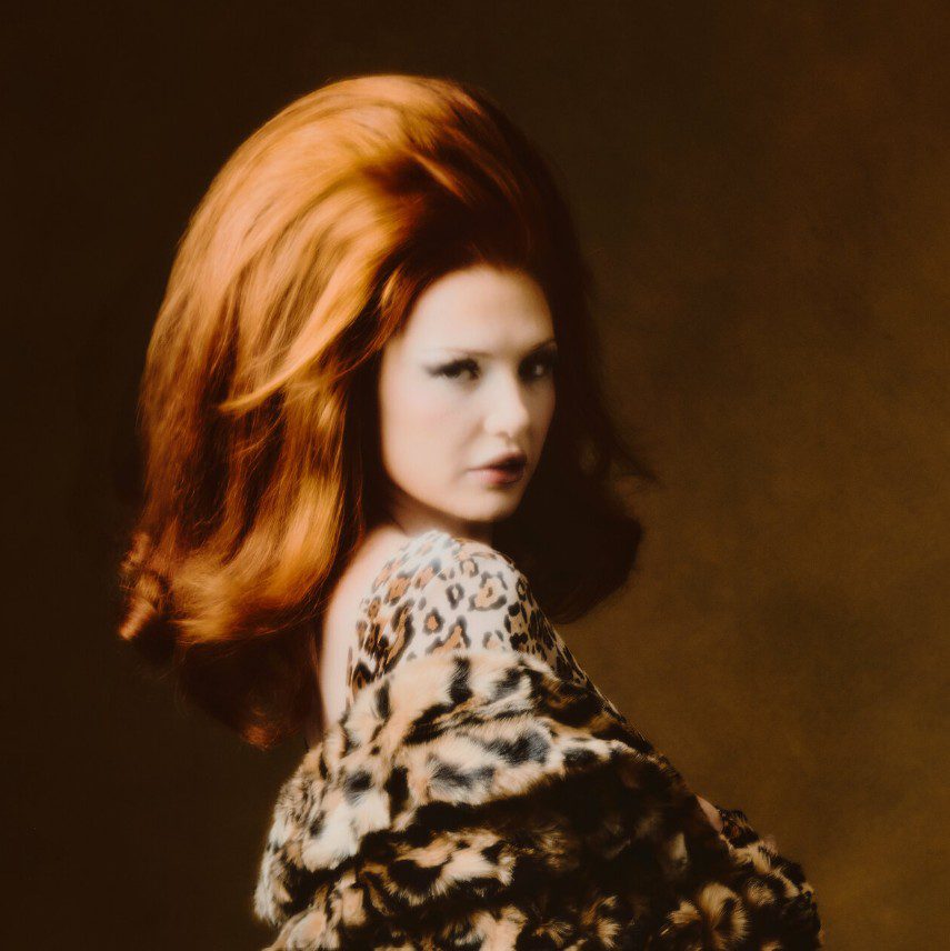 Slightly out of focus image of Queenie, looking over her shoulder at the camera against a brown background. Single cover for Vivid.