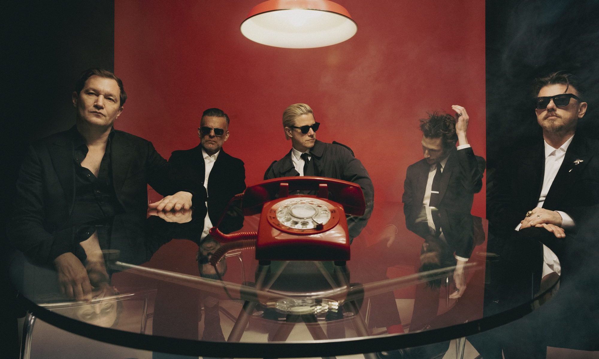 Interpol press image - band dressed in black suits around a glass table with large red rotary phone on it.