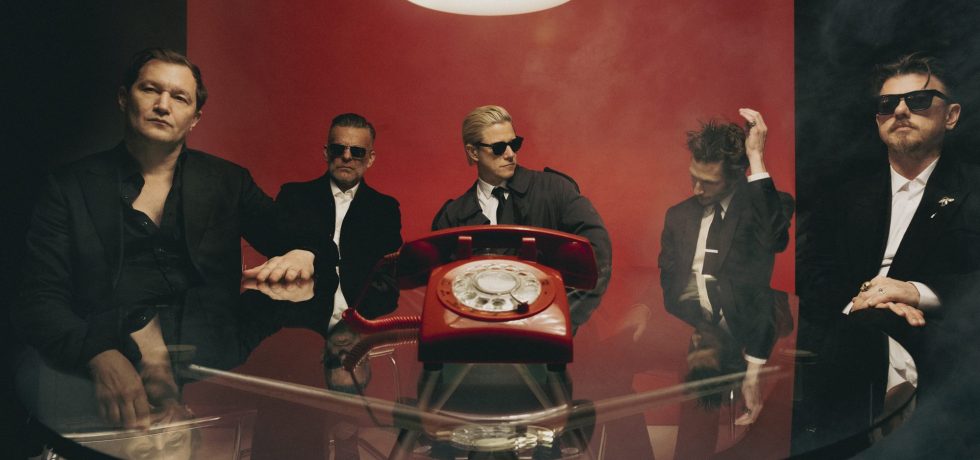 Interpol press image - band dressed in black suits around a glass table with large red rotary phone on it.