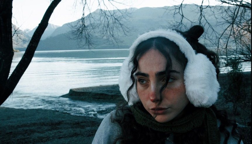 Image of Charli Lucas in front of a wintery looking lake wearing ear muffs. Single Cover for Drinking About It