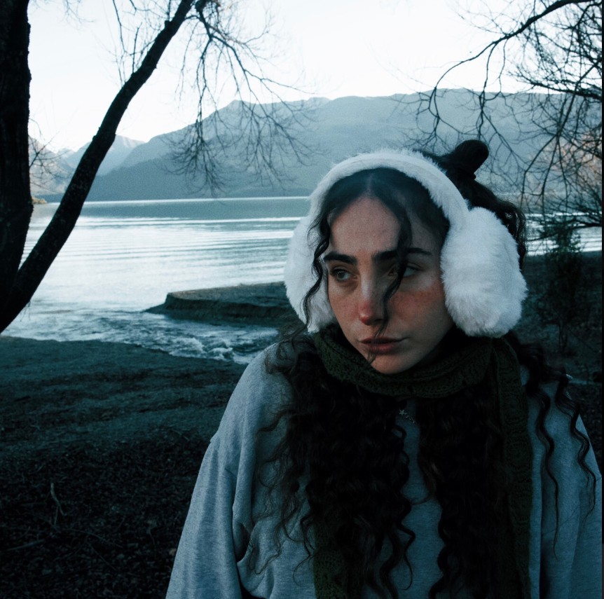 Image of Charli Lucas in front of a wintery looking lake wearing ear muffs. Single Cover for Drinking About It